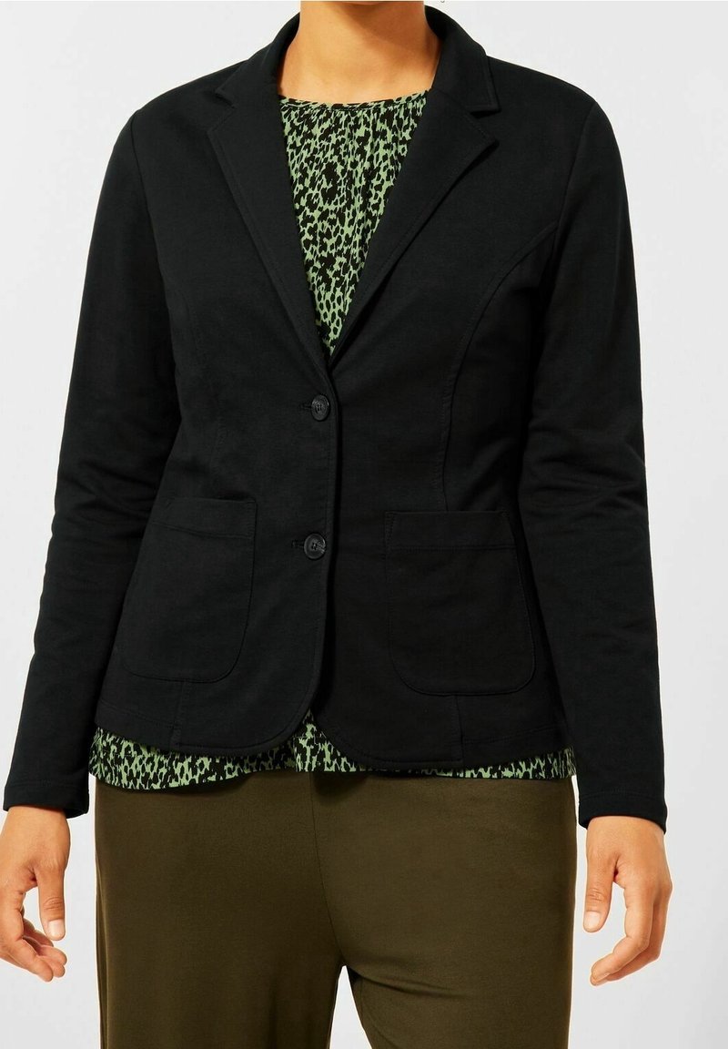 Personne portant un blazer noir ajusté à deux boutons sur un haut à motifs verts et noirs et un pantalon vert olive.