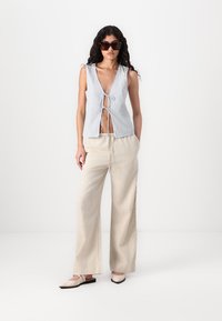 Haut sans manches à rayures avec fermeture à nouer à l'avant, associé à un pantalon beige à jambes larges. Le mannequin porte des lunettes de soleil noires et des mocassins plats.