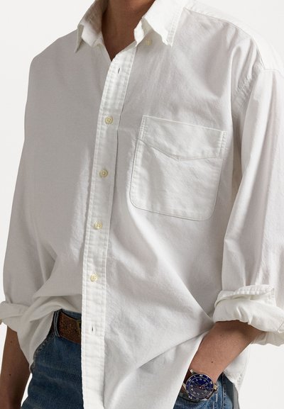 Camisa blanca de botones con un solo bolsillo en el pecho, mangas arremangadas y una textura sutil; combinada con jeans azules y un reloj de pulsera.