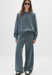 Preveliki teget dukserica s rebrastim manžetama i odgovarajućim širokim hlačama, izrađeni od mekanog materijala. Model kombinira ovu odjeću s tamnim naočalama i crnim cipelama.