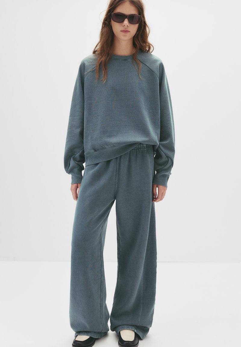 Preveliki teget dukserica s rebrastim manžetama i odgovarajućim širokim hlačama, izrađeni od mekanog materijala. Model kombinira ovu odjeću s tamnim naočalama i crnim cipelama.