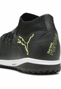 Zapatilla deportiva negra con diseño de punto, lados texturizados y suela de goma. Presenta un logo verde y el texto "JUEGA LIBRE FUTURO" en el talón.