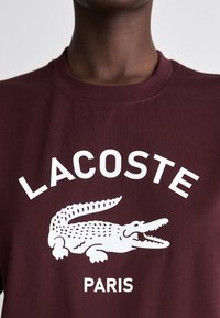 Burgundi medvilnės marškinėliai, kurie išsiskiria ryškiu baltu užrašu "LACOSTE" ir krokodilo logotipu, apvaliu kaklu, lygia tekstūra, minimalizmo stiliumi.