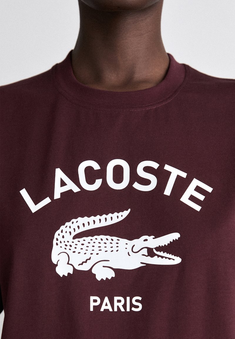 Camiseta de algodón burdeos con destacado texto en blanco "LACOSTE" y logo de cocodrilo, diseño de cuello redondo, textura suave, estilo minimalista.