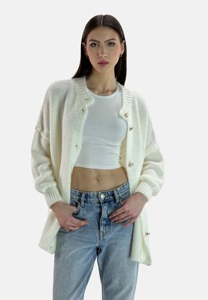 Cardigan lavorato a maglia color crema con una vestibilità comoda, scollo rotondo e bottoni dorati. Indossato sopra un top bianco aderente e jeans in denim chiaro a vita alta.
