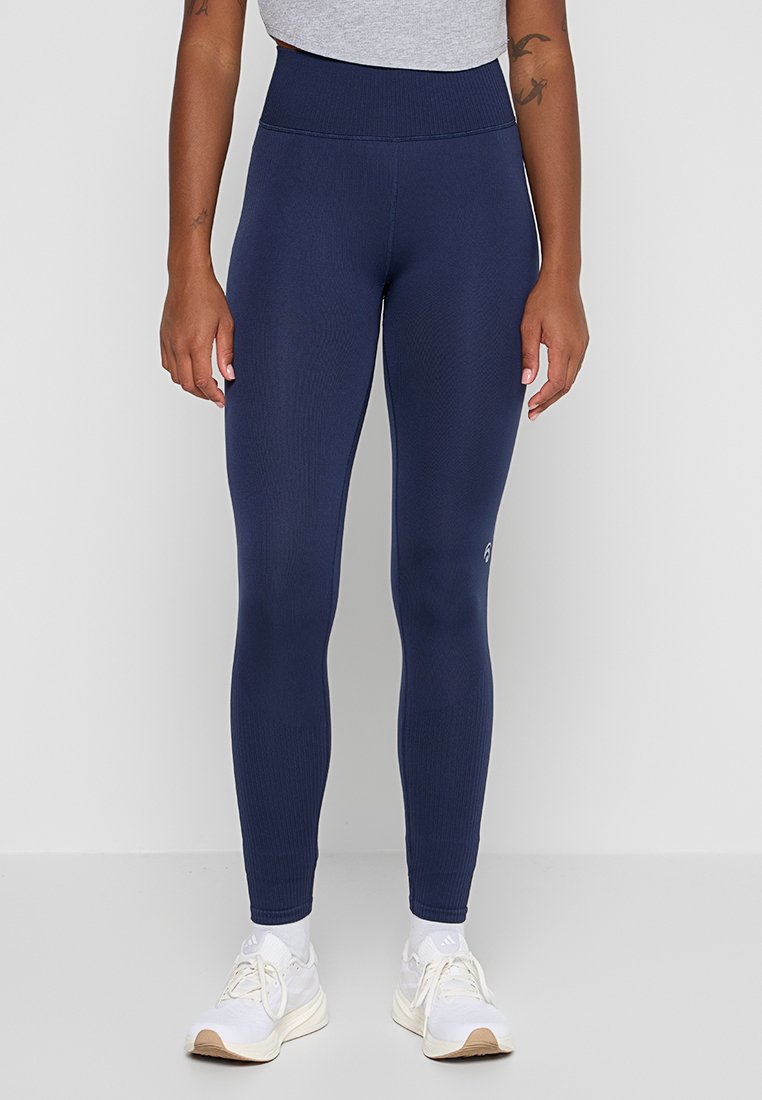 Frau mit marineblauen High-Waist-Leggings und weißen Sneakers, die vor einem einfarbigen hellgrauen Hintergrund steht.