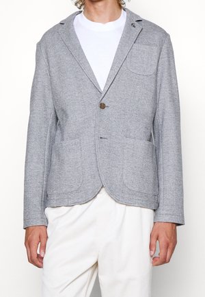 Homme portant une veste décontractée gris clair avec trois poches plaquées et un seul bouton marron, un T-shirt blanc et un pantalon crème.