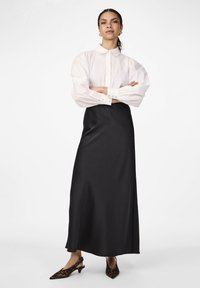 Camicia bianca con bottoni e maniche a sbuffo, abbinata a una gonna lunga nera in satin. Gli accessori includono orecchini a cerchio dorati e décolleté con stampa leopardata.