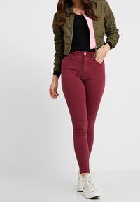 Donna con giacca bomber cropped verde oliva, top nero, jeans skinny bordeaux e sneakers alte bianche, in piedi su sfondo bianco.