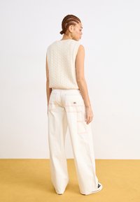 Beige Strickpullover mit Zopfmuster, kombiniert mit weißen weiten Hosen mit rosa Nähten und mehreren Taschen, von hinten gesehen.