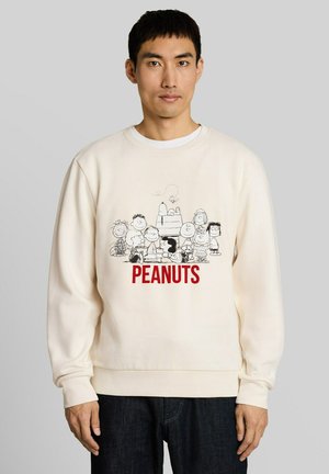 Mężczyzna noszący jasny kremowy sweatshirt z czarno-białymi postaciami z komiksu Peanuts i słowem "PEANUTS" na przodzie napisanym na czerwono.