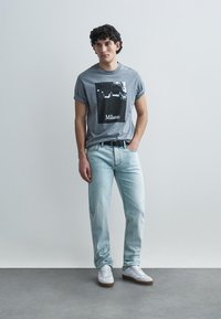 T-shirt cinzenta de algodão com uma estampa fotográfica a preto e branco rotulada como "Milano", combinada com jeans de ganga azul claro e sapatilhas brancas.