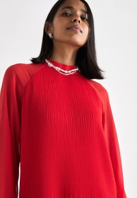 Crvena plisirana majica s dugim prozirnim rukavima i okruglim izrezom. Ima slojevitu teksturu i srebrni lančić kao naglasak.