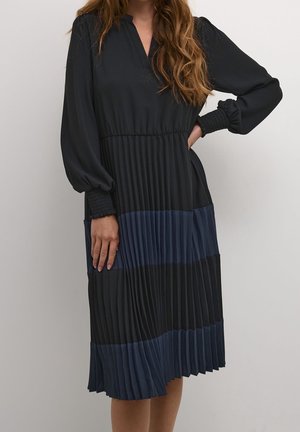 Vrouw in een zwarte, knielange jurk met lange mouwen, smockmanchetten en een geplooide rok met afwisselende zwarte en donkerblauwe horizontale strepen.