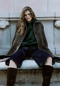 Abrigo de tweed marrón sobre una sudadera verde, combinado con pantalones cortos de mezclilla azul marino. La modelo lleva botas de ante marrón hasta la rodilla, con cabello largo y ondulado.