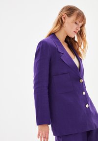 Blazer in lino viola con un design sartorial, tasca sul petto singola e bottoni dorati. Vestibilità strutturata con maniche lunghe e rever a punte.