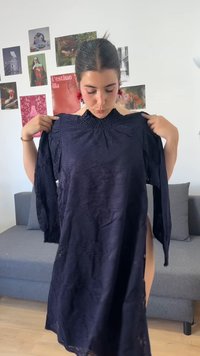 Γυναίκα με σκούρα μαλλιά κρατά ένα μακρυμάνικο σκούρο μπλε φόρεμα μπροστά της, στέκεται σε ένα δωμάτιο με γκρι καναπέ και έργα τέχνης στον τοίχο πίσω της.