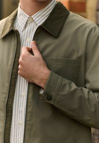 Chaqueta de color verde oliva con cuello de pana, puños abotonados y un bolsillo delantero, superpuesta sobre una camisa blanca de rayas. Textura suave.
