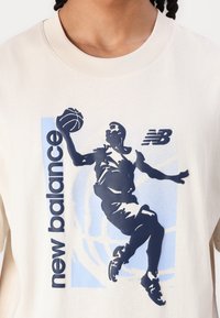 Kremna trenerka z grafiko košarkarja v modri in temno modri barvi ter napisom "new balance". Rebrast ovratnik in sproščen kroj.