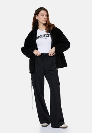 Kobieta ubrana w czarną, puszystą kurtkę na zamek błyskawiczny, białą koszulkę z napisem "Brooklyn", czarne szerokie spodnie typu cargo oraz szare sneakersy, stoi z rękami na linii pasa.
