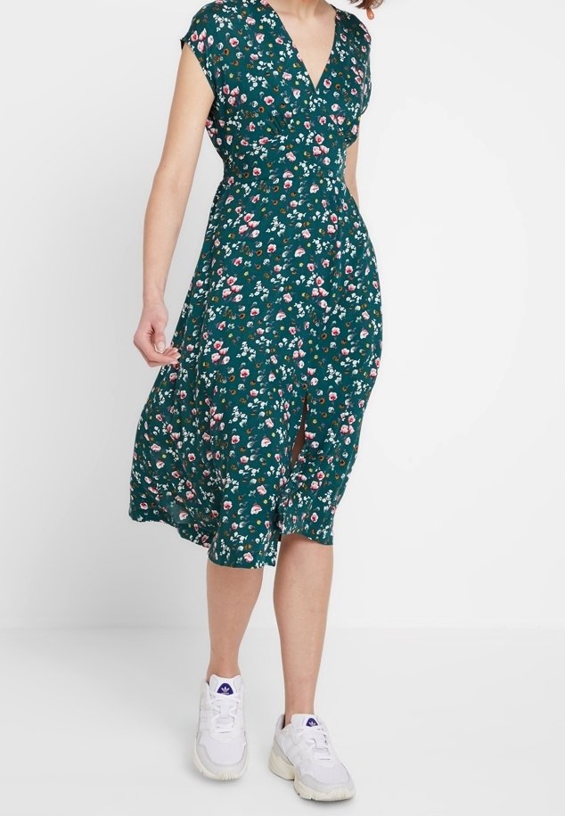 Femme portant une robe midi verte à fleurs avec une fente devant et des baskets blanches, marchant sur un fond blanc uni.