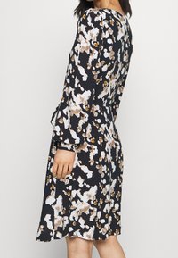 Šaty s květinovým vzorem a dlouhými rukávy, mají černý základ a bílé, hnědé a oranžové abstraktní designy. Hladká tkanina, délka po kolena.