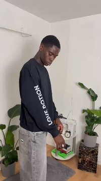 Sweatshirt preto com "LOVE POTION" impresso em branco ao longo da manga esquerda. Usado com calças de ganga cinzentas, em meio a plantas e decoração interiores.