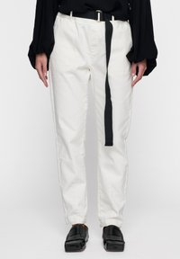 Pantaloni bianchi in cotone con una vestibilità rilassata, dotati di una cintura in tessuto nera, tasche laterali e una superficie liscia. Abbinati a scarpe nere slip-on.
