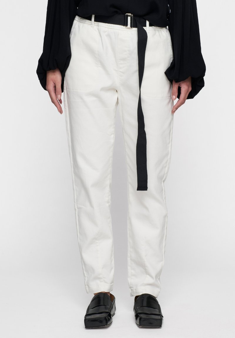 Witte katoenen broek met een relaxte pasvorm, voorzien van een zwarte stoffen riem, zijzakken en een gladde textuur. Gecombineerd met zwarte instapschoenen.