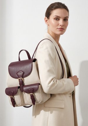 Donna con cappotto beige che indossa uno zaino beige in tela con cinghie e patta in pelle marrone scuro, in piedi su sfondo chiaro uniforme.