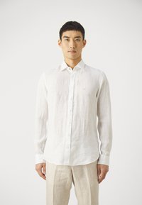 Camisa de lino blanca de manga larga con cuello button-down, botones en el frente y un sutil logo bordado en el pecho. Textura suave, corte relajado.