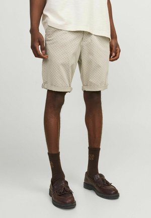 Homme portant un short beige retroussé avec de petits pois, des chaussettes marron foncé avec texte, et des mocassins à pampilles marron foncé, debout devant un fond uni.