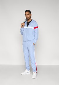 Άνδρας με ανοιχτόγαλανη φόρμα με κόκκινες και λευκές ρίγες, κρατώντας ρακέτα τένις στον ώμο, στέκεται μπροστά σε απλό φόντο.