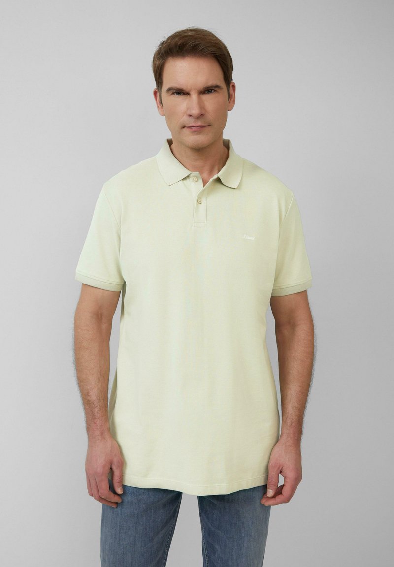 Bleke gele polo shirt met een klassiek ontwerp, korte mouwen en een knoopsluiting met drie knopen. De stof heeft een subtiele textuur. Draagt met blauwe jeans.