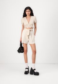 Top court crème avec manches bouffantes et dentelle, associé à un short beige taille haute et des chaussures plates noires. Sac noir à motifs.