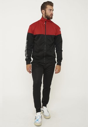Uomo che indossa una giacca rossa e nera con zip, pantaloni neri e sneakers bianche, in piedi e guarda di lato su sfondo bianco.