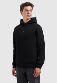 Jonge man in een eenvoudige zwarte hoodie met de capuchon omlaag en donkere jeans, kijkend naar links tegen een eenvoudige lichtgrijze achtergrond.