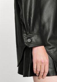 Černý kožený oděv se strukturovanými rameny a zapínáním na manžetách. Látka má hladkou texturu s mírným leskem, který ukazuje čisté linie.