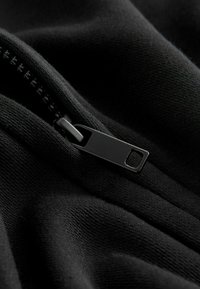 Tissu noir avec une fermeture éclair visible. La tirette de la fermeture éclair est noire mate, avec un trou rectangulaire. La texture semble douce et flexible.