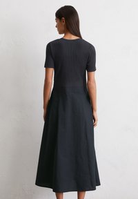 A woman stands with her back to the viewer, wearing a fitted black top and a flowy black skirt, against a textured light wall.