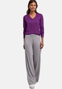 Fioletowa bluzka z długim rękawem i dekoltem w serek oraz jasnoszare szerokie spodnie z miękkiego materiału. Modelka stoi, ubrana w białe trampki, ręce luźno wzdłuż ciała.