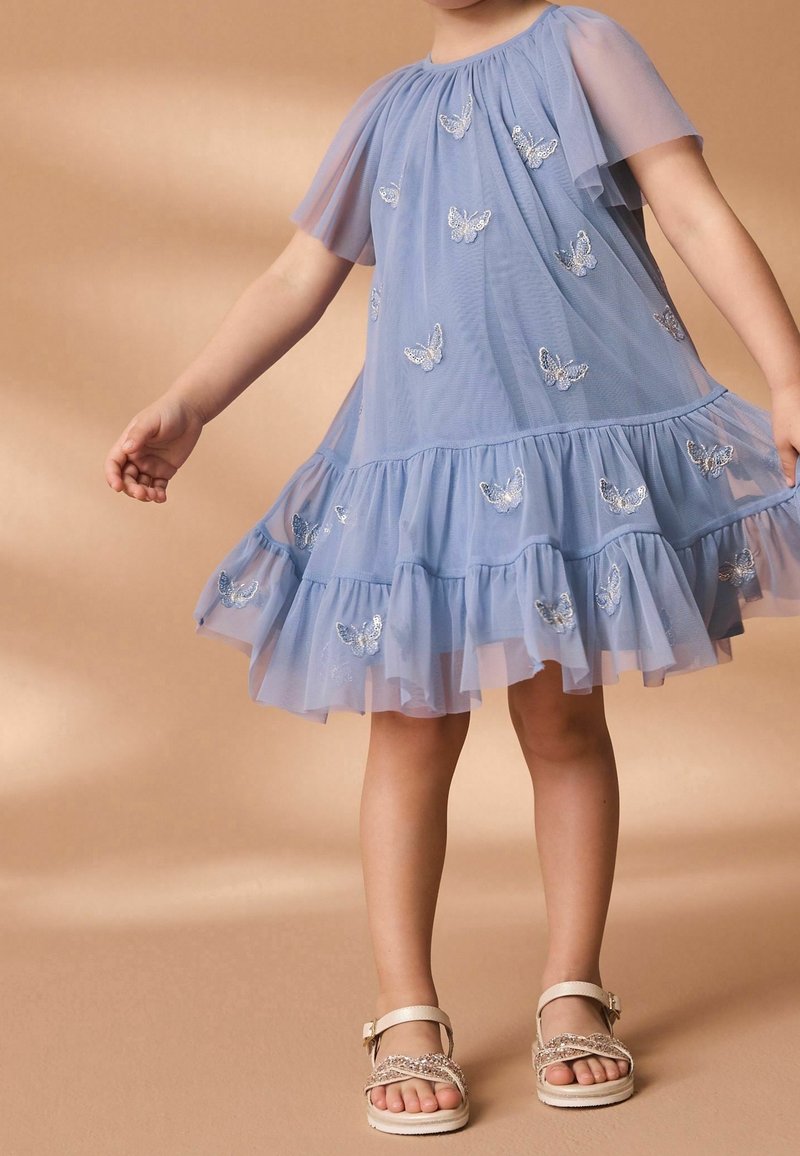 Enfant portant une robe en tulle bleu avec une broderie papillon argentée et des sandales scintillantes, debout devant un fond neutre.