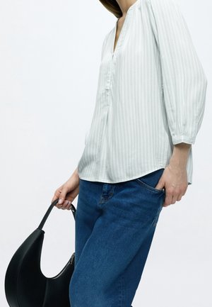 Personne portant un chemisier blanc à manches longues et un jean bleu, portant un sac à main noir avec une poignée courbée.