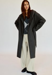 Lange, grijze wollen jas met een grote, puntige kraag, gedragen over een gebreid patroontrui en brede beige broeken. Zwarte, platte schoenen maken de look compleet.