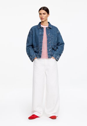 Kobieta w niebieskiej kurtce dżinsowej na czerwono-białej pasiastce, białe spodnie o szerokich nogawkach i czerwone buty, stojąca z rękami w kieszeniach.