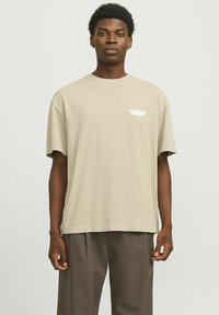 Beige bomulds T-shirt med en afslappet pasform, korte ærmer og hvidt trykt tekst på brystet. Parret med løse mørkebrune bukser.