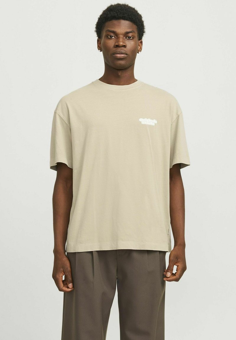 Beige bomulds T-shirt med en afslappet pasform, korte ærmer og hvidt trykt tekst på brystet. Parret med løse mørkebrune bukser.