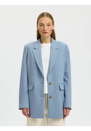 Kvinne med rett blondt hår som har på seg en oversized lys blå blazer, hvit skjorte og beige bukser, står mot en ensfarget bakgrunn.