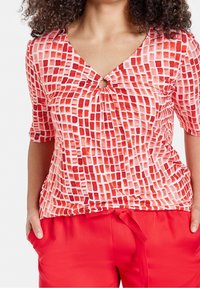 Nainen, joka on pukeutunut punavalkoiseen ruudulliseen puseroon, jossa on pieni avaus edessä, ja tummanpunaiseen housupariin, pitäen käsiään taskuissa.