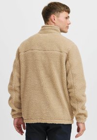 Chaqueta de forro polar beige con un exterior texturizado y esponjoso. Cuenta con un collar alto y mangas largas, diseñada con un corte holgado y una costura sencilla.